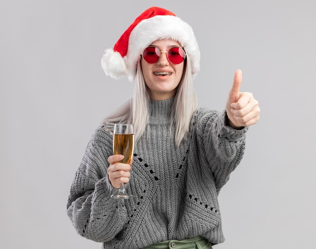 Jovem loira com um suéter de inverno e um chapéu de papai noel segurando uma taça de champanhe, parecendo feliz e positiva, sorrindo mostrando os polegares para cima