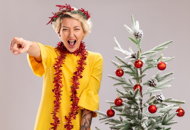 Jovem loira confiante com coroa de flores de natal e guirlanda de ouropel em volta do pescoço, em pé perto de uma árvore de natal decorada, olhando apontando em linha reta gritando isolado na parede branca