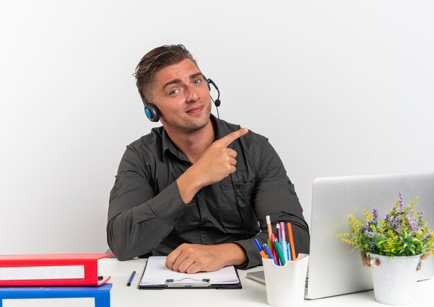 Jovem loira confiante, trabalhador de escritório, usando fones de ouvido, sentado na mesa com ferramentas de escritório, usando laptop aponta ao lado isolado no fundo branco com espaço de cópia