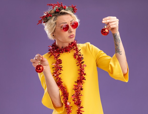 Jovem loira confusa com uma coroa de flores de Natal e uma guirlanda de ouropel em volta do pescoço com óculos segurando enfeites de natal, olhando para um deles isolado no fundo roxo