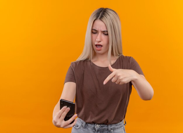 Jovem loira confusa segurando um telefone celular apontando e olhando para ele em um espaço laranja isolado com espaço de cópia