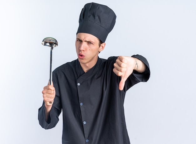 Jovem loira cozinheira carrancuda com uniforme de chef e boné segurando uma concha, olhando para a câmera mostrando o polegar isolado na parede branca