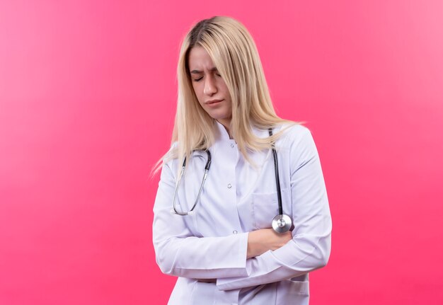 Jovem loira de médico triste usando estetoscópio e vestido de médico, cruzando as mãos sobre fundo rosa