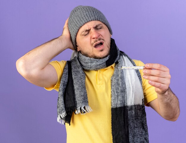 Foto grátis jovem loira doente com dor e chapéu de inverno e lenço coloca a mão na cabeça olhando para o termômetro isolado na parede roxa