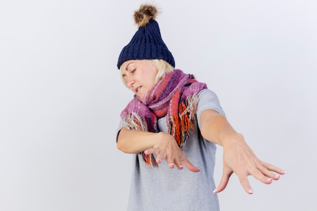 Jovem loira doente, irritada, usando chapéu de inverno e cachecol com as mãos isoladas na parede branca Foto gratuita