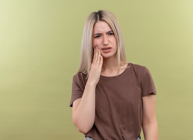 Jovem loira dolorida colocando a mão na bochecha e sofrendo de dor de dente em um espaço verde isolado com espaço de cópia