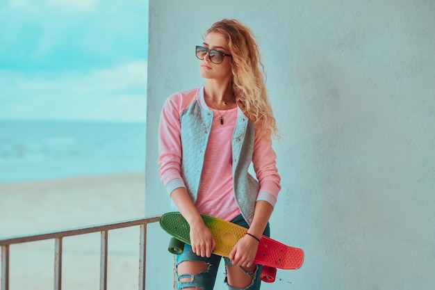 Jovem loira em óculos de sol está posando para o fotógrafo enquanto segura seu skate.