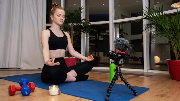 Jovem loira em roupas esportivas meditando em um tapete de ioga olhando para a gravação