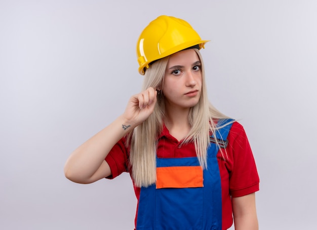 Jovem loira engenheira construtora de uniforme colocando o dedo na orelha olhando para uma parede branca