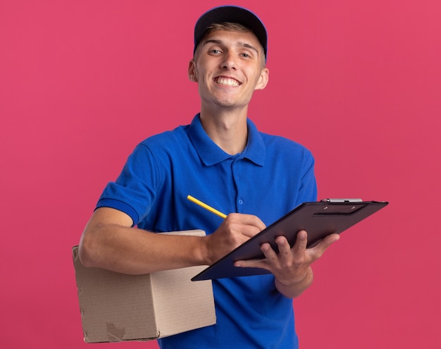 Foto grátis jovem loira entregadora sorridente segurando caixa de papelão e escrevendo na prancheta com lápis isolado na parede rosa com espaço de cópia