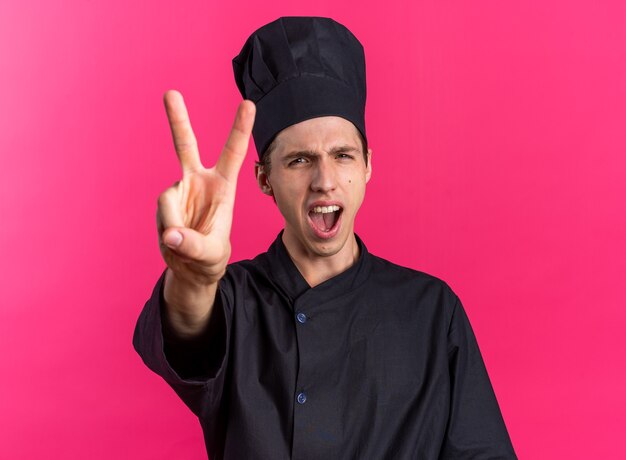 Jovem loira estrita cozinheiro com uniforme de chef e boné olhando para a câmera gritando, mostrando dois com a mão isolada na parede rosa