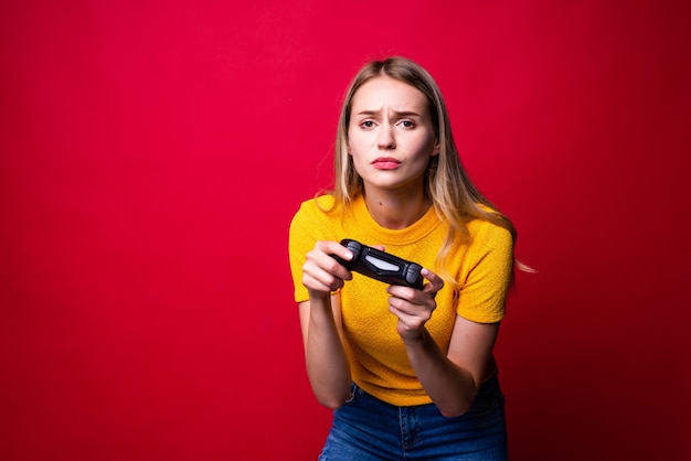 Jovem loira gamer usando gamepad jogando videogame isolado na parede vermelha