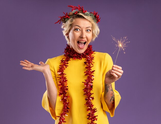 Jovem loira impressionada com coroa de flores de Natal e guirlanda de ouropel em volta do pescoço segurando diamante de feriado, olhando para a câmera, mostrando a mão vazia isolada no fundo roxo