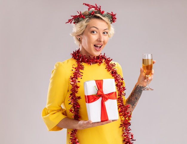 Jovem loira impressionada com coroa de flores de Natal e guirlanda de ouropel em volta do pescoço, segurando uma taça de champanhe e um pacote de presente, olhando para a câmera, isolada no fundo branco