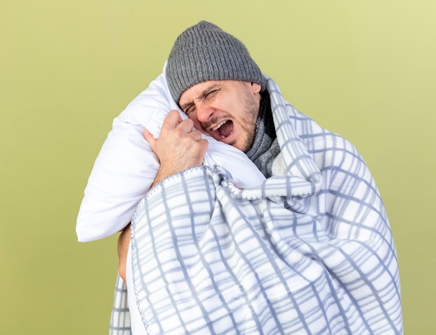 Jovem loira irritada com um homem doente usando um chapéu de inverno envolto em um travesseiro de abraços xadrez isolado na parede verde oliva