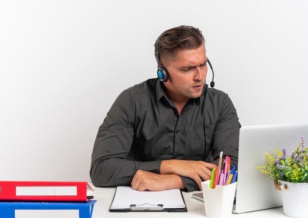 Jovem loira irritada, trabalhador de escritório, usando fones de ouvido, sentado na mesa com ferramentas de escritório, olhando para um laptop isolado no fundo branco com espaço de cópia