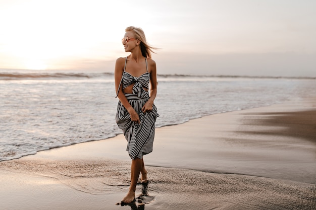 Jovem loira legal em um vestido listrado elegante e óculos de sol rosa caminhar na praia, sorrir e curtir o pôr do sol