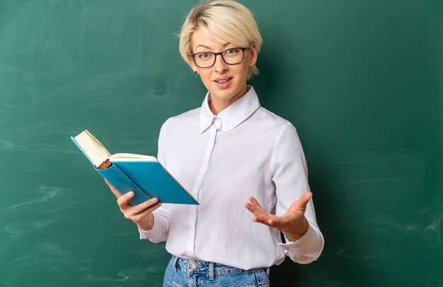 Jovem loira professora animada usando óculos em uma sala de aula em frente ao quadro-negro, segurando o livro, mostrando a mão vazia com espaço de cópia