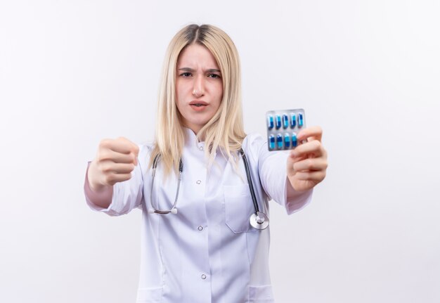 Jovem loira rigorosa, usando estetoscópio e bata médica, segurando comprimidos e segurando o punho fechado para a câmera em fundo branco isolado