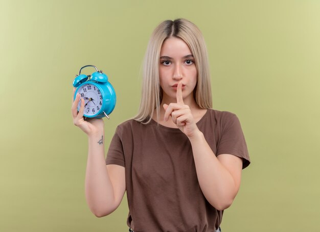 Jovem loira segurando um despertador gesticulando silêncio na parede verde isolada