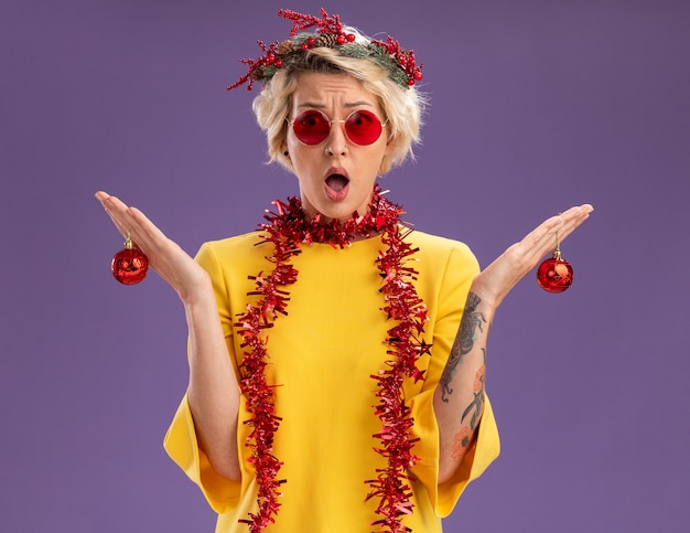 Foto grátis jovem loira sem noção usando coroa de flores de natal e guirlanda de ouropel em volta do pescoço segurando enfeites de natal e parecendo isolada na parede roxa