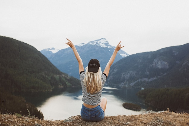 Jovem loira sentada na falésia apreciando a vista das montanhas e do lago