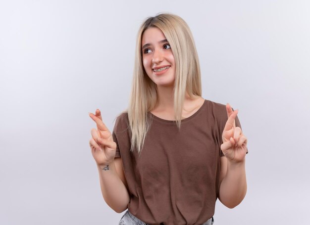 Jovem loira sorridente com aparelho dentário fazendo gestos de dedos cruzados em um espaço em branco isolado com espaço de cópia