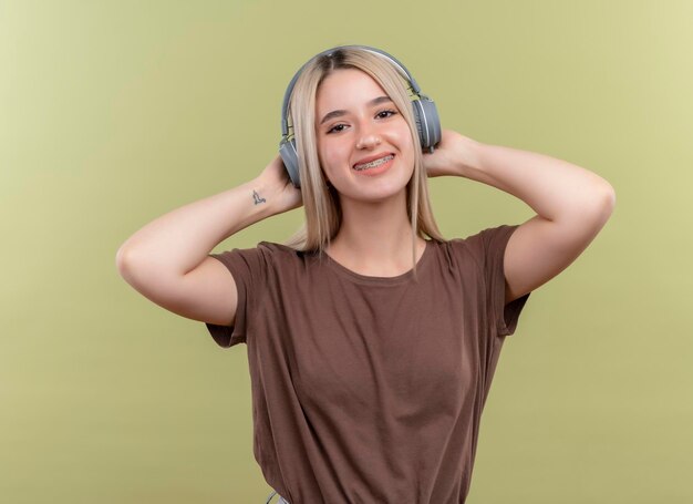 Jovem loira sorridente com aparelho dentário usando fones de ouvido e colocando as mãos atrás da cabeça em um espaço verde isolado