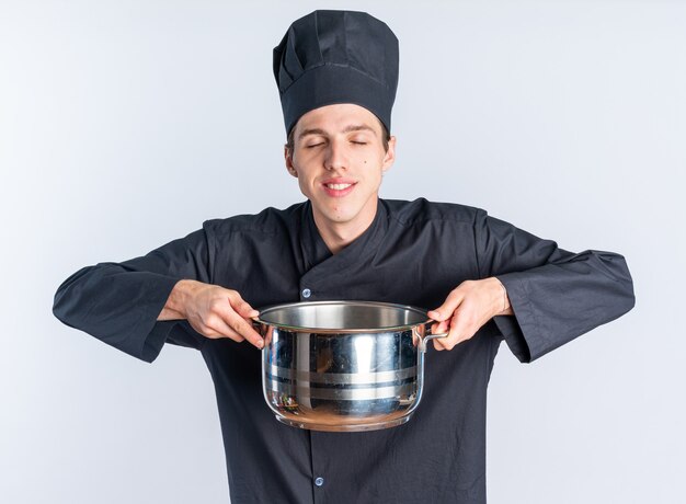 Foto grátis jovem loira sorridente com uniforme de chef e boné segurando a panela com os olhos fechados, isolada na parede branca