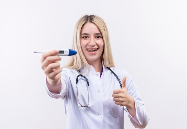Jovem loira sorridente de médico usando estetoscópio e bata médica com aparelho dentário segurando o termômetro na câmera e o polegar para cima no fundo branco isolado
