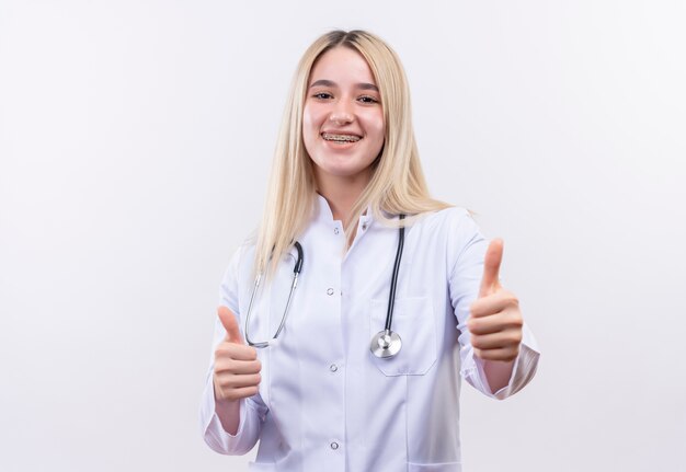 Jovem loira sorridente de médico usando estetoscópio e bata médica em aparelho dentário com os polegares para cima em fundo branco isolado