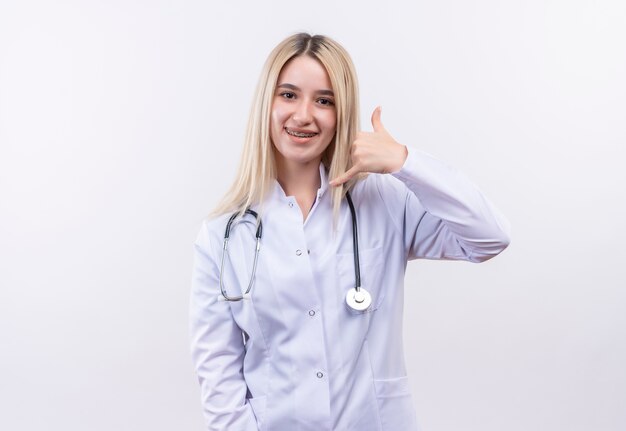 Jovem loira sorridente de médico usando estetoscópio e bata médica em aparelho dentário, mostrando gesto de chamada em fundo branco isolado