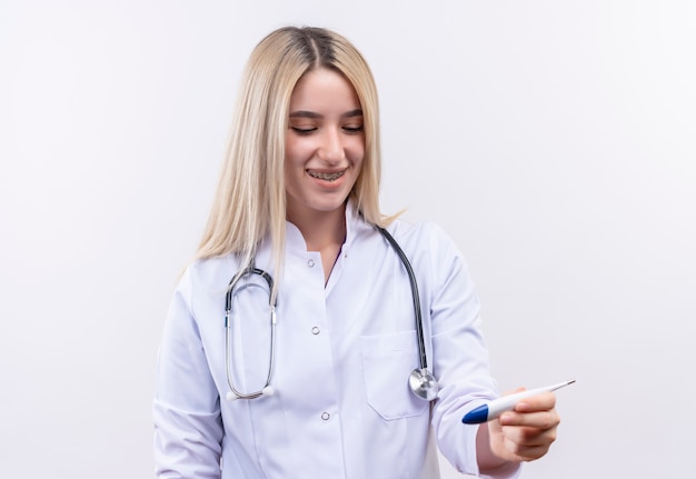 Jovem loira sorridente de médico usando estetoscópio e bata médica em aparelho dentário, olhando o termômetro na mão em fundo branco isolado