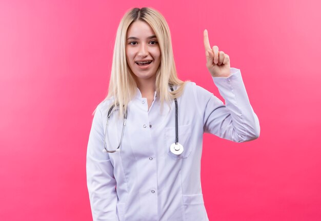 Jovem loira sorridente de médico usando estetoscópio em bata médica e aparelho dentário aponta para cima em fundo rosa isolado