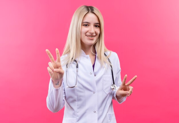 Jovem loira sorridente de médico usando um estetoscópio com bata médica e aparelho dentário, mostrando um gesto de paz com as duas mãos no fundo rosa isolado