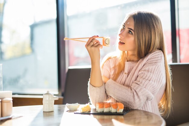 Jovem loira sorridente e ensolarada com um suéter branco comendo sushi no almoço em um pequeno café