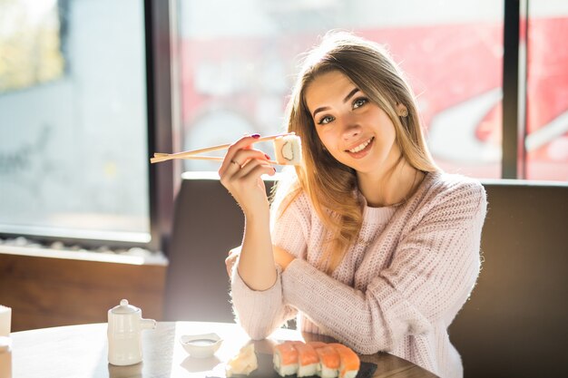 Jovem loira sorridente e ensolarada com um suéter branco comendo sushi no almoço em um pequeno café