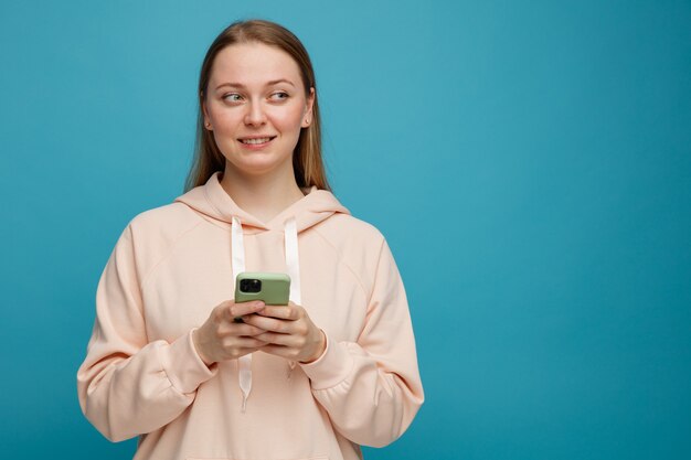 Jovem loira sorridente segurando um celular e olhando para o lado