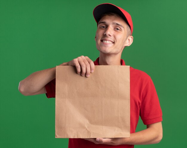 Jovem loira sorridente segurando um pacote de papel isolado em uma parede verde com espaço de cópia
