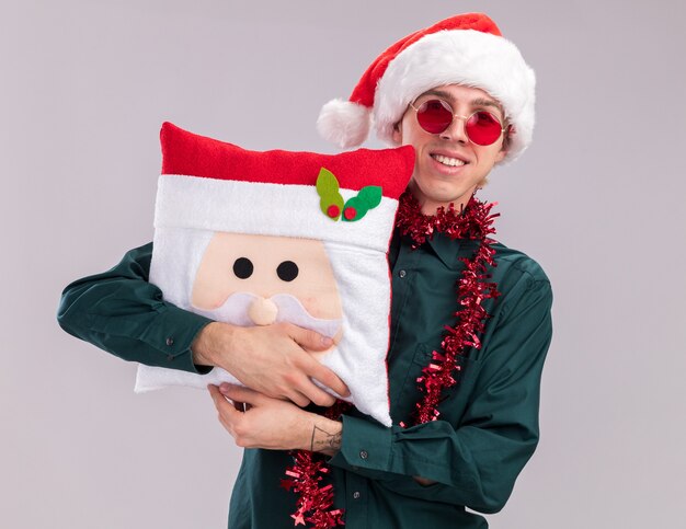 Jovem loira sorridente usando chapéu de Papai Noel e óculos com guirlanda de ouropel no pescoço abraçando a almofada de Papai Noel, olhando para a câmera isolada no fundo branco