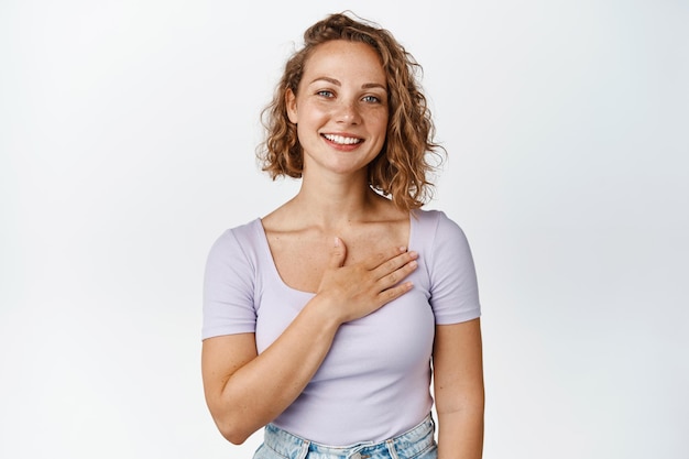 Jovem loira sorrindo, segurando a mão no coração e parecendo lisonjeada, diga obrigado, de pé contra o fundo branco