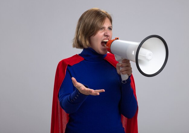 Jovem loira super-heroína furiosa com capa vermelha gritando no alto-falante, olhando em linha reta, mostrando a mão vazia isolada na parede branca com espaço de cópia