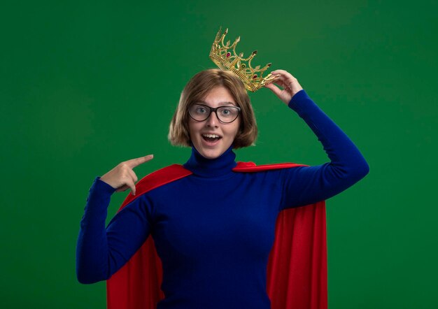 Jovem loira super-heroína impressionada com uma capa vermelha usando óculos e uma coroa, agarrando uma coroa, olhando para a frente e apontando para si mesma, isolada na parede verde