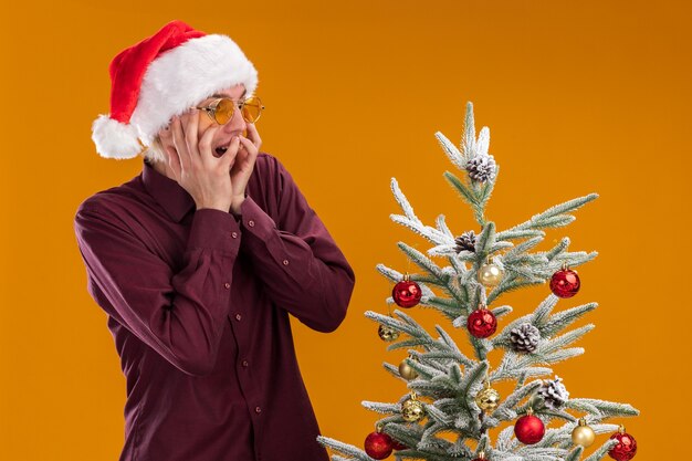 Jovem loiro animado com chapéu de Papai Noel e óculos em pé perto da árvore de Natal decorada, com as mãos no rosto olhando para baixo, isolado em um fundo laranja