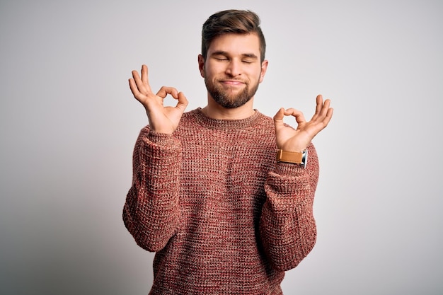 Foto grátis jovem loiro com barba e olhos azuis vestindo suéter casual sobre fundo branco relaxado e sorrindo com os olhos fechados fazendo gesto de meditação com os dedos conceito de yoga