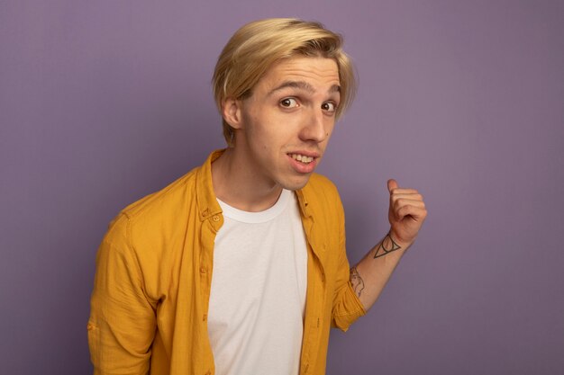 Jovem loiro impressionado com uma camiseta amarela apontando para trás, isolado em roxo com espaço de cópia