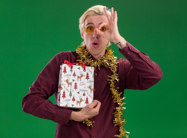 Jovem loiro impressionado usando óculos com guirlanda de ouropel no pescoço segurando uma sacola de presente de Natal fazendo gesto de olhar isolado na parede verde