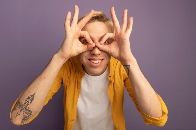 Jovem loiro sorridente usando uma camiseta amarela e mostrando um gesto de olhar isolado no roxo