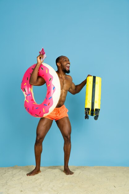 Jovem louco e feliz descansando com um anel de praia como um donut e uma bolsa no espaço azul