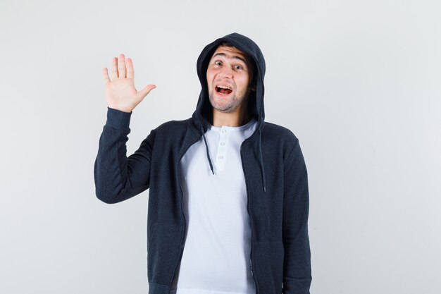 Jovem macho acenando a mão para saudação em t-shirt, jaqueta e olhando alegre, vista frontal.